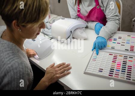 Concetto: Cura personale, manicure. Manicurista che lavora. Giovane ragazza che fa lavoro di unghia sulle mani di donna adulta. Scelta da vernice acrilica. Grafico a colori. Palet Foto Stock