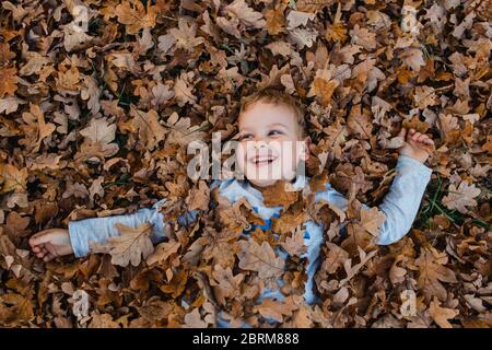 Ragazzo giovane che giace sulle foglie autunnali a terra. Carino bambino coperto di foglie di autunno marrone secco sorridente e guardando via. Foto Stock