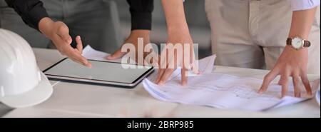 Primo piano di due ingegneri che consultano il loro progetto con tablet e documenti mock-up su un tavolo bianco nella sala riunioni Foto Stock