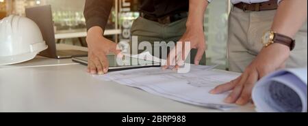 Un'immagine ritagliata di due ingegneri che lavorano insieme a un tablet e a una documentazione su un tavolo bianco nella sala riunioni Foto Stock