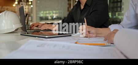 Un'immagine ritagliata di due ingegneri che lavorano insieme a BluePrint, tablet digitale e notebook sulla scrivania dell'ufficio Foto Stock