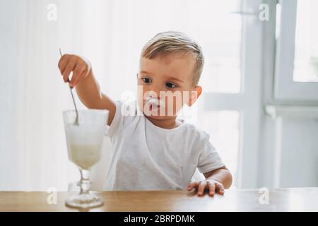 Piccolo bambino concentrato di charme che mangia yogurt dal vetro nella luminosa cucina di casa Foto Stock
