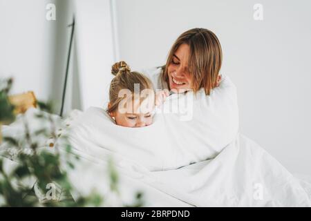 Carino ragazza toddler lungo capelli lisci occhi grigi grandi guardando la macchina fotografica divertirsi con la madre a letto soggiorno a casa Foto Stock