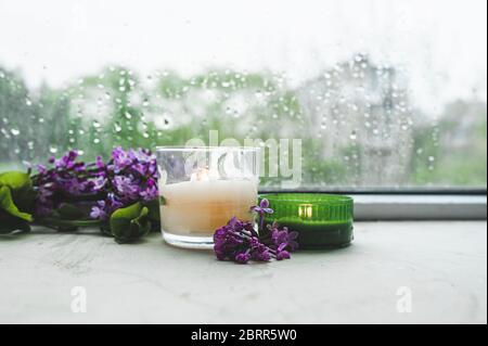 bruciando due candele bianche e verdi con rami di fresco porpora fiore lilla sprig sullo sfondo di vetro bagnato in gocce e gocce dal vento Foto Stock