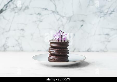 tre biscotti di brownie al cioccolato accatastati su un piatto di porcellana bianca con un fiore di lilla sulla parte superiore contro uno sfondo bianco di marmo Foto Stock