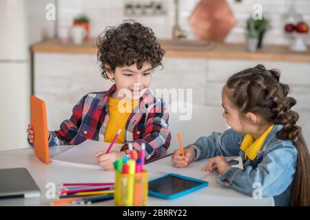 Ragazzo curly e ragazza dai capelli scuri seduto a tavola con le tavolette, facendo i loro compiti, discutendo qualcosa Foto Stock