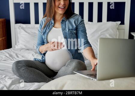 Donna incinta irriconoscibile che usa il laptop a letto Foto Stock