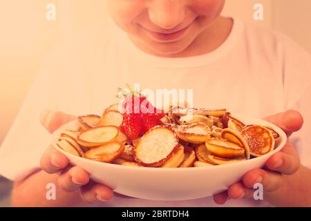 Happy boy con ciotola bianca con mini pancake sani alla moda, cereali e fragole. Nessuna immagine del volto, concetto di famiglia. Sana colazione vegana senza Foto Stock