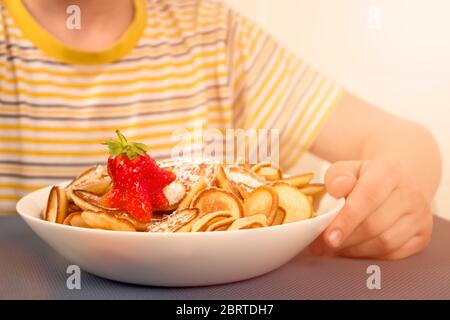 Bambino ragazzo mangiare sano trendy mini pancake cereali e fragole rosse da ciotola bianca. Nessuna immagine del volto, concetto di famiglia. Sana colazione vegana Foto Stock