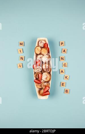 Posa piatta verticale composizione minima con mini frittelle di cereali con fragole su piastra bianca e lettere di legno su sfondo blu con spazio per la copia Foto Stock