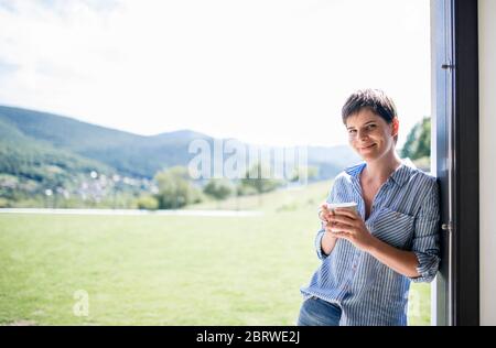 Vista frontale della giovane donna con caffè in piedi accanto alla porta del patio a casa. Foto Stock