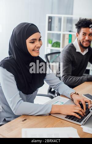 Fuoco selettivo di sorridente donna d'affari araba usando il laptop vicino collega afroamericano in ufficio Foto Stock