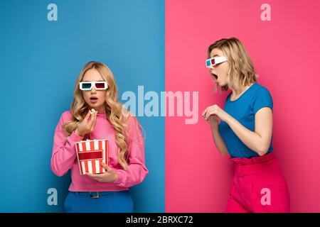 Scioccata ragazza bionda in occhiali 3d guardando sorella mangiare popcorn su sfondo blu e rosa Foto Stock