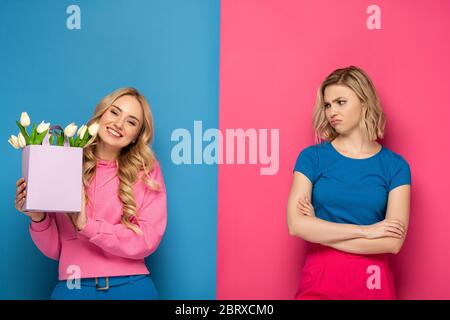 Ragazza sorridente che tiene bouquet vicino envy sorella bionda su sfondo blu e rosa Foto Stock