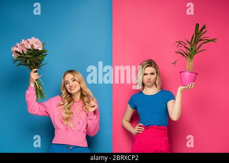 Ragazza sorridente che tiene bouquet vicino sorella offesa con pianta su sfondo rosa e blu Foto Stock