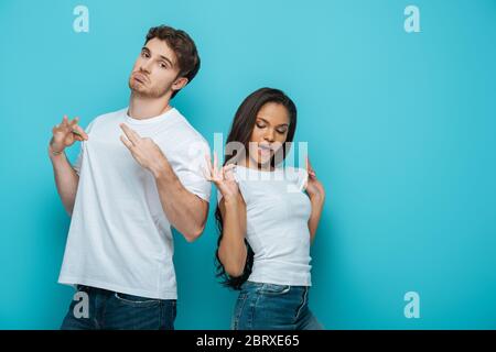 uomo scettico e ragazza afro-americana flirty che tocca la t-shirt bianca su sfondo blu Foto Stock