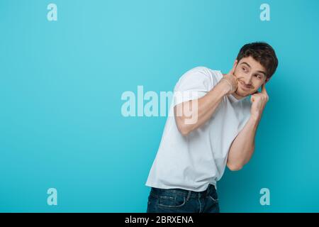 dispiaciuto giovane uomo che ostruiva le orecchie con le dita mentre guardò via su sfondo blu Foto Stock