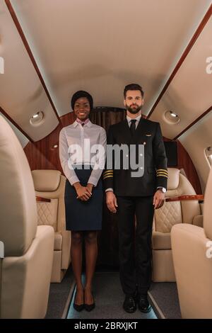 bel pilota sicuro e sorridente, afroamericano, stewardess, guardando la telecamera in aereo privato Foto Stock