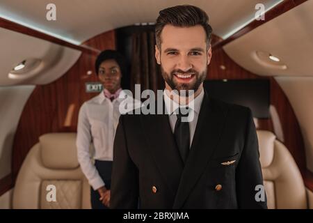 fuoco selettivo del bel pilota sorridente alla telecamera vicino alla stewardess afroamericana Foto Stock