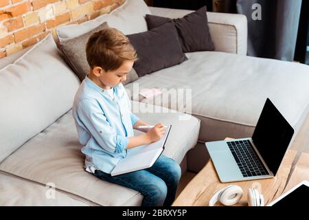 adorabile ragazzo che scrive in notebook vicino a gadget con schermo vuoto mentre e-learning a casa Foto Stock