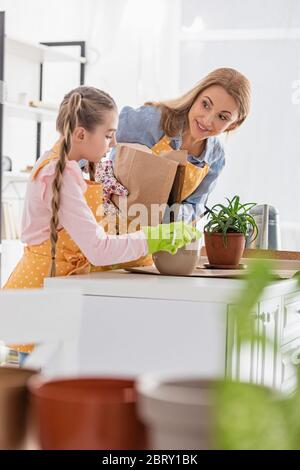 Fuoco selettivo di madre sorprendo che mette terra al vaso di fiori e guardando la figlia con rastrello vicino a tavola in cucina Foto Stock