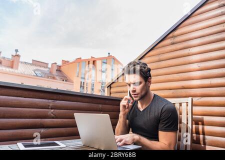 uomo giovane e serio che usa il computer portatile e parla con lo smartphone mentre si siede sul balcone Foto Stock