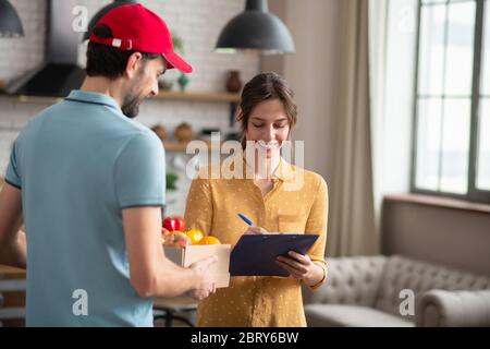 Cliente femminile dai capelli scuri che firma la lettera di vettura dopo aver ricevuto i generi alimentari e sorridendo Foto Stock