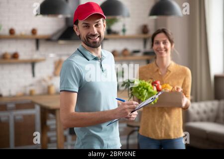 Giovane Courier maschio in un cappello rosso che tiene generi alimentari e sorridente Foto Stock