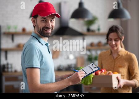 Giovane Courier maschio in un cappello rosso che tiene generi alimentari e sorridendo piacevolmente Foto Stock