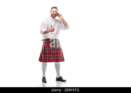 Felice uomo scozzese rosso in kilt rosso che parla su smartphone e mostra il pollice su sfondo bianco Foto Stock