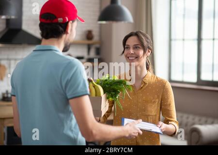 Cliente dai capelli scuri che riceve generi alimentari consegnati dal corriere e sorridente Foto Stock