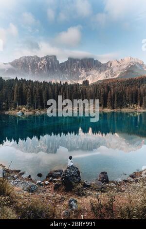 Indietro veduta di una ragazza bionda felice contro il cielo blu sul Lago di Karersee, Dolomiti, Italia. Donna spensierata con abito floreale e un cappello gode la vista lago su Foto Stock