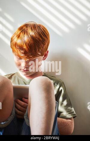 Ragazzo con capelli rossi seduto sul pavimento in una stanza soleggiata, tenendo un tablet digitale. Foto Stock