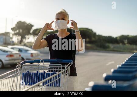 Donna con carrello di shopping che mette la maschera protettiva sul viso appena prima di entrare in un negozio Foto Stock