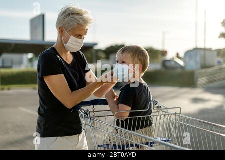 Madre che aiuta suo figlio seduto in un'automobile di shopping a indossare una maschera protettiva prima di entrare in un negozio. Foto Stock