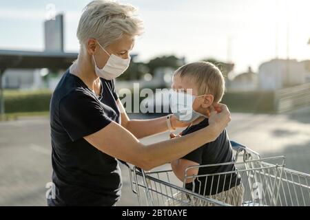 Madre che aiuta suo figlio seduto in un'automobile di shopping a indossare una maschera protettiva prima di entrare in un negozio. Foto Stock