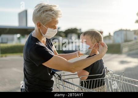 Madre che aiuta suo figlio seduto in un'automobile di shopping a indossare una maschera protettiva prima di entrare in un negozio. Foto Stock