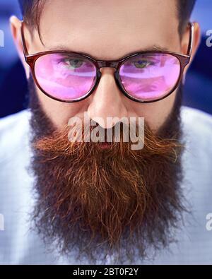 Uomo bearded in occhiali con screen reflection che lavora su computer in ufficio IT, seduto alla scrivania di scrittura codice, lavorando su un progetto in software Foto Stock