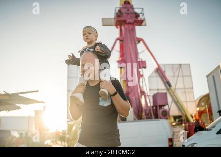 Felice padre con il suo piccolo figlio piggybacked in un parco di divertimenti Foto Stock