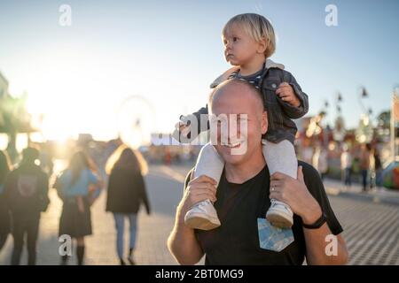 Felice padre con il suo piccolo figlio piggybacked in un parco di divertimenti Foto Stock