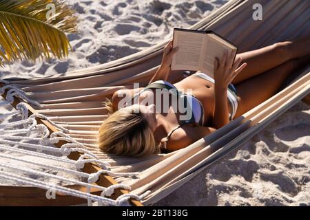 Donna caucasica che si stese su un'amaca e che legge un libro in spiaggia. Foto Stock