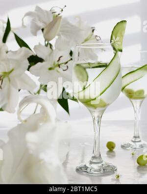 Un alto bicchiere con gin e tonico servito con striscia di cetriolo su sfondo di fiori bianchi. Foto Stock
