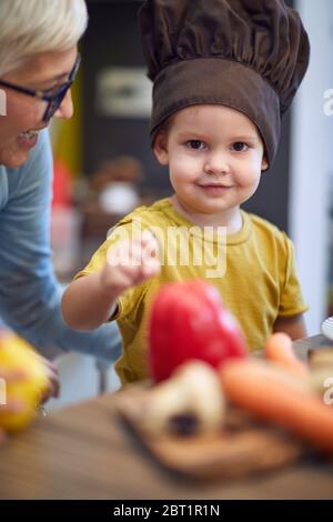 Bel ragazzo in cappello da cuoco che passa il tempo felice in cucina con la nonna. Foto Stock