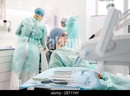 Giovane donna in hap monouso e accappatoio seduta in sedia e in attesa di dentista e assistente che si prepara per la procedura di guarigione in clinica Foto Stock