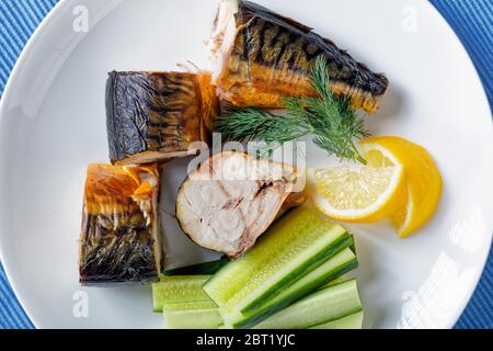 Piatto antipasto: Fette di sgombro affumicato caldo con senape di digione, cetrioli freschi, fette di aneto e limone, posate dorate su un panno blu da tavola su una w Foto Stock