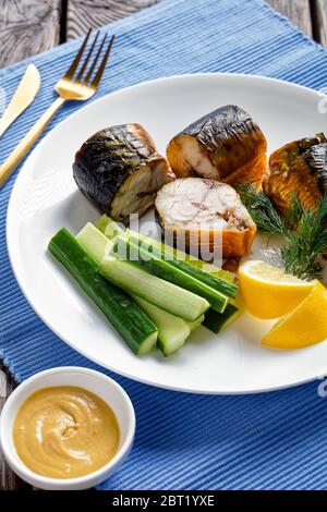 Sgombri dell'atlantico pescati selvatici affumicati caldi serviti su un piatto bianco con senape di digione, cetrioli freschi, aneto e spicchi di limone, posate dorate su un blu Foto Stock