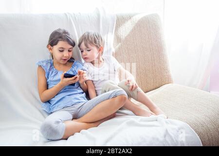 I bambini a casa si siedono sul divano e giocano con il tablet. Fratelli e sorelle carini hanno un buon tempo a casa. I bambini giocano un gioco su un telefono cellulare Foto Stock
