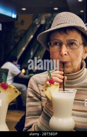 Bella vecchia donna di stile che beve un cocktail dolce al bar. Vecchia donna caucasica in cappello e bicchieri sorseggiando drink in ristorante godendo la vita. Buon ma Foto Stock