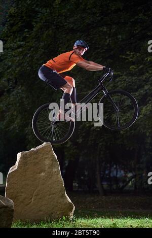 Vista frontale del ciclista che guarda nella telecamera durante un salto in bicicletta. Atleta che mantiene l'equilibrio sulla ruota posteriore della mountain bike sportiva. Concetto di stile di vita attivo. Foto Stock