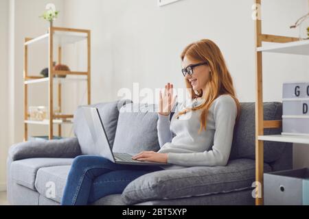Felice giovane femmina in occhiali sorridente e salutare l'amico mentre si siede sul divano e si effettua una videochiamata a casa Foto Stock